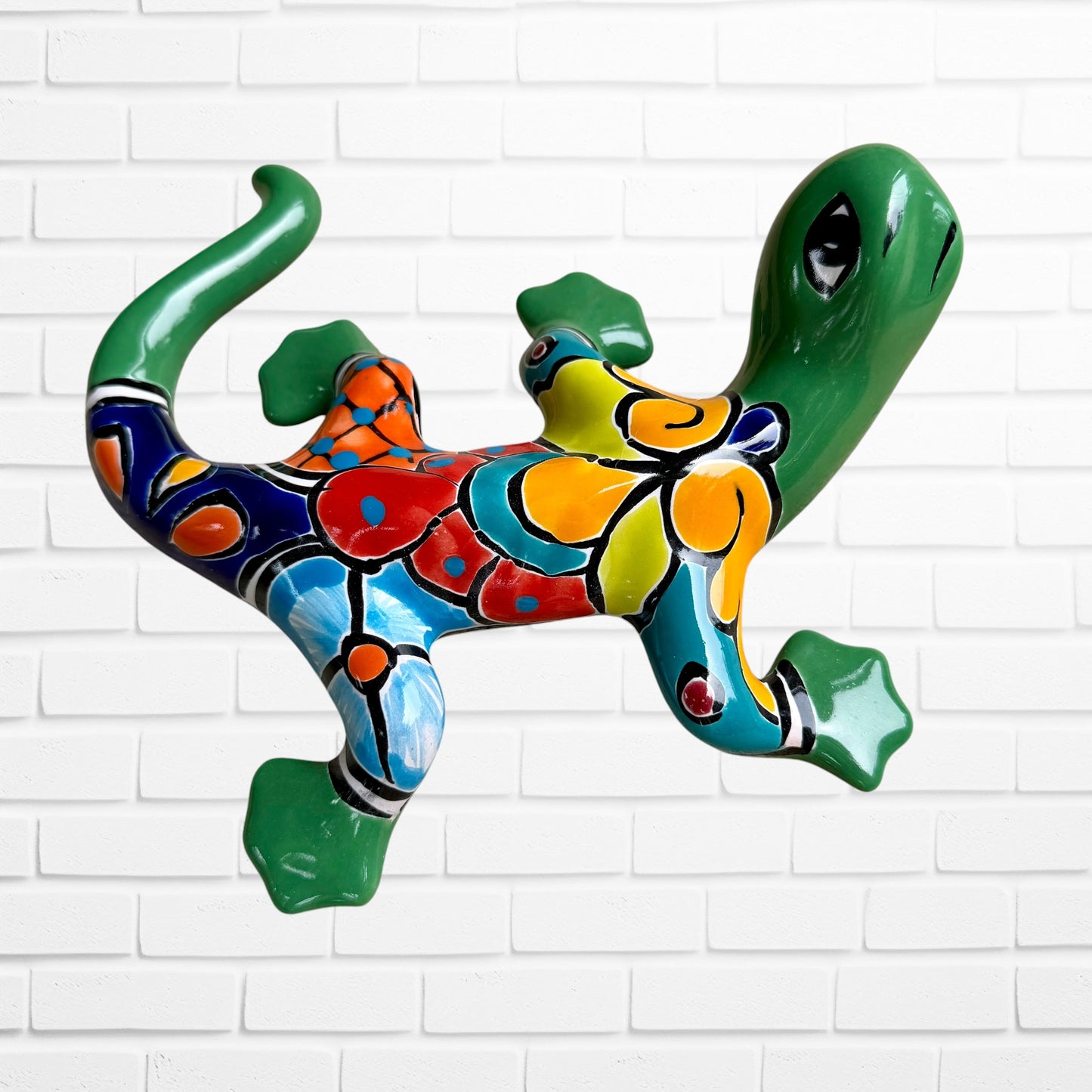 A colorful hand-painted ceramic lizard wall sculpture mounted on a white brick wall. The lizard features a vibrant mosaic pattern with green, blue, red, yellow, and orange sections back side view.