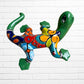 A colorful hand-painted ceramic lizard wall sculpture mounted on a white brick wall. The lizard features a vibrant mosaic pattern with green, blue, red, yellow, and orange sections back side view.