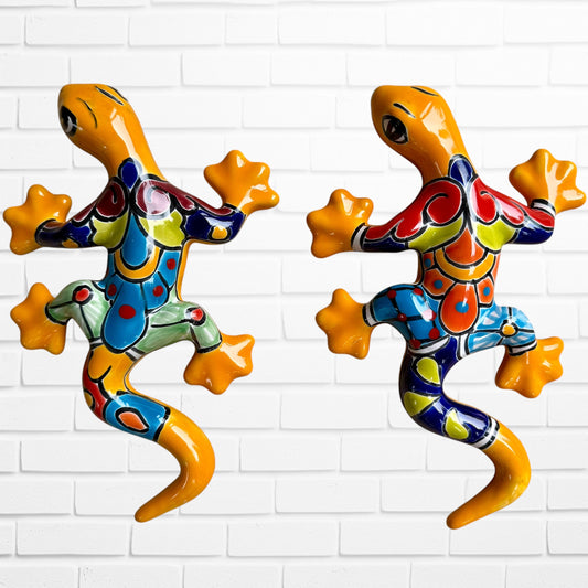 Two colorful hand-painted ceramic gecko wall sculptures in a vibrant Mexican folk art style, displayed side-by-side against a white brick wall.