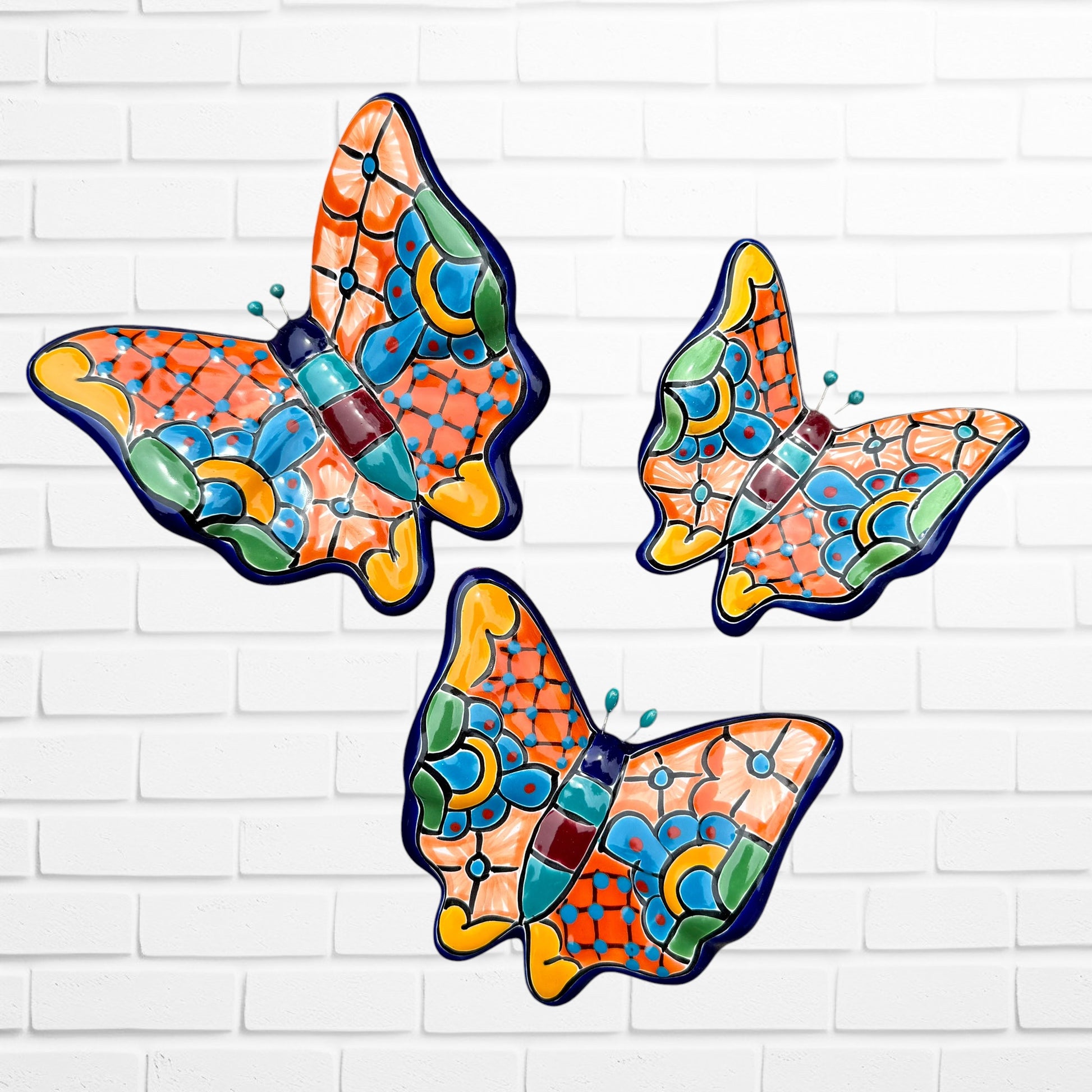 Three colorful ceramic Talavera butterfly wall art pieces with orange, blue, green, yellow, and purple mosaic patterns, displayed against a white brick wall.