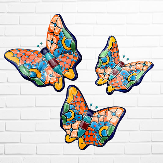 Three colorful ceramic Talavera butterfly wall art pieces with orange, blue, green, yellow, and purple mosaic patterns, displayed against a white brick wall.