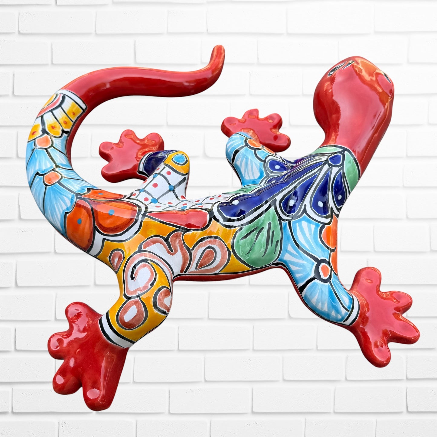 A colorful Talavera ceramic wall salamander lizard art piece with a glossy finish, featuring a red head and tail with a patterned body, displayed against a white brick wall background, right side body view.