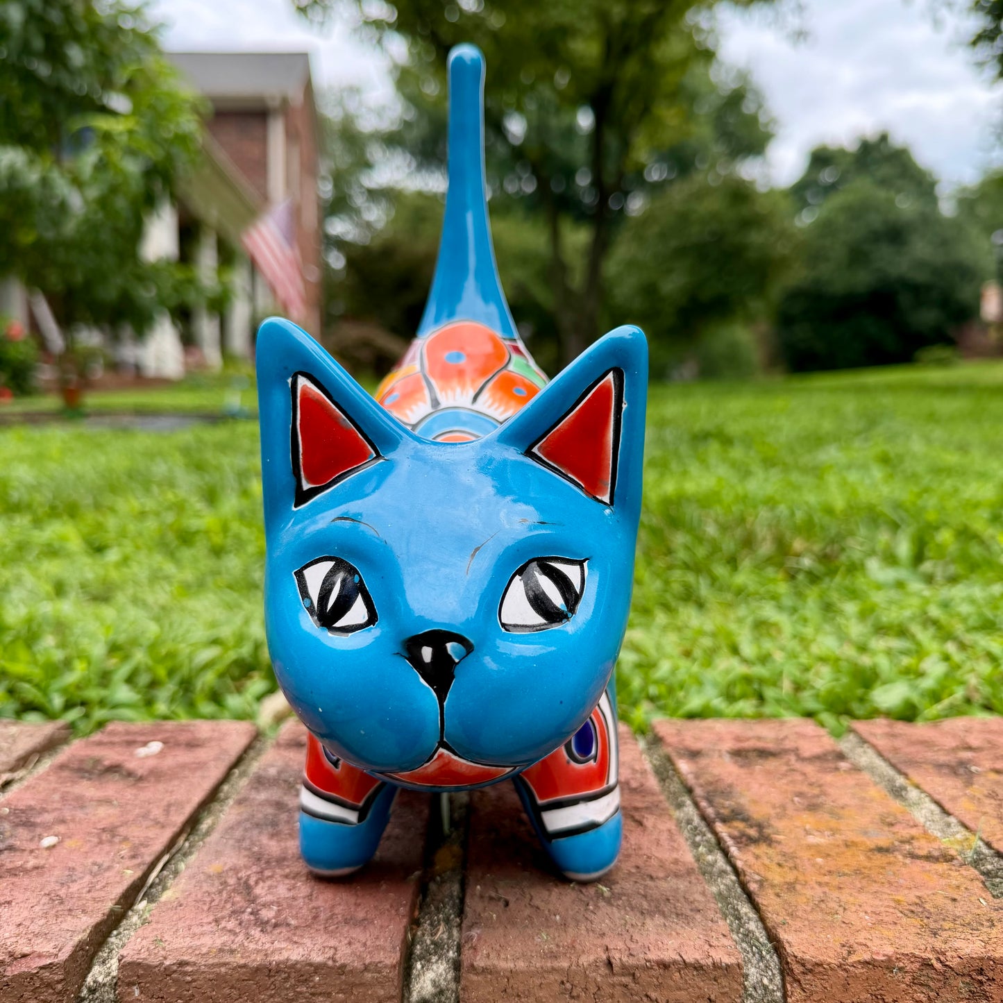 A glossy talavera ceramic cat figurine decorated with vibrant hand-painted patterns in turquoise, yellow, green and red, standing on a brick path with a green lawn and a house in the blurred background front face view.