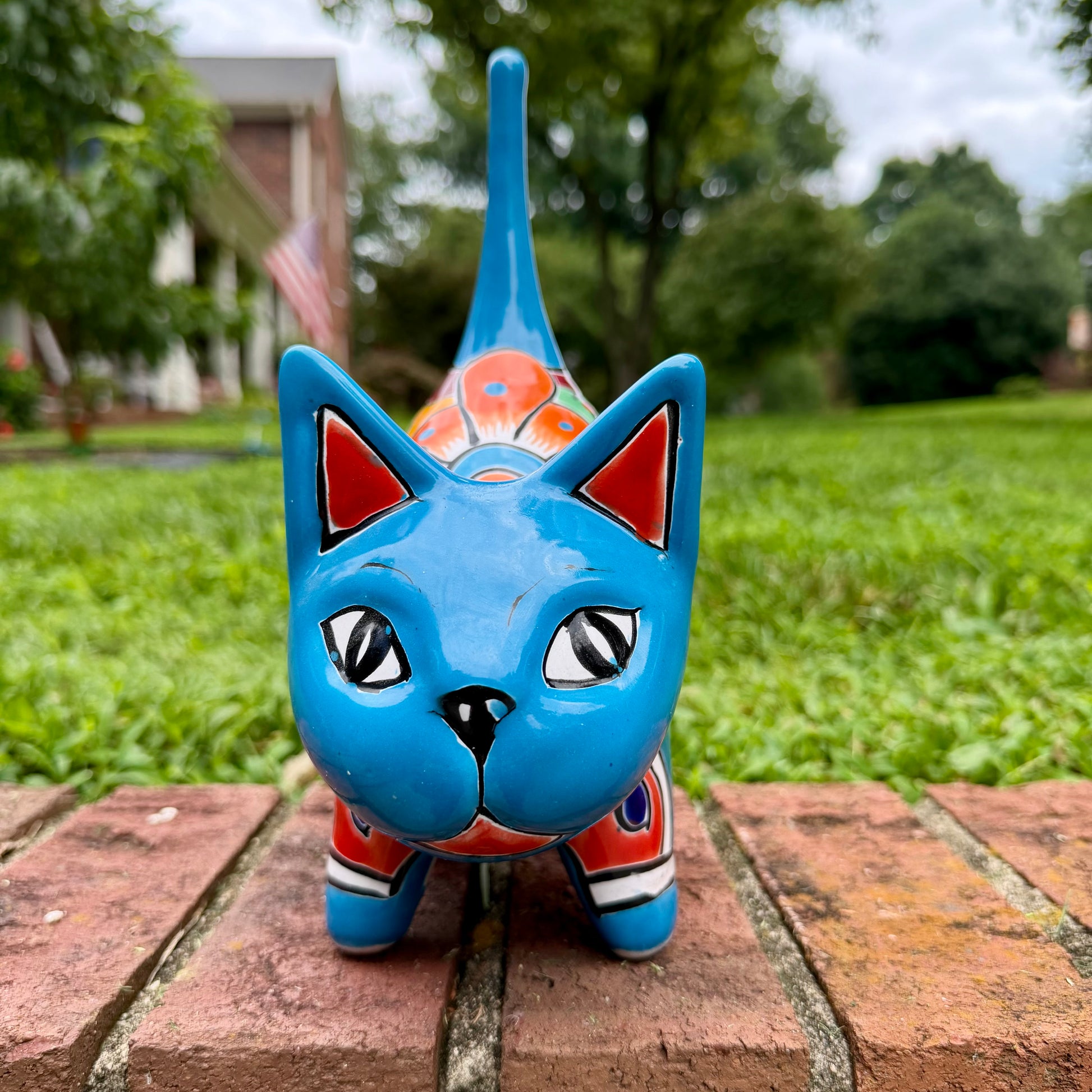 A glossy talavera ceramic cat figurine decorated with vibrant hand-painted patterns in turquoise, yellow, green and red, standing on a brick path with a green lawn and a house in the blurred background front face view.