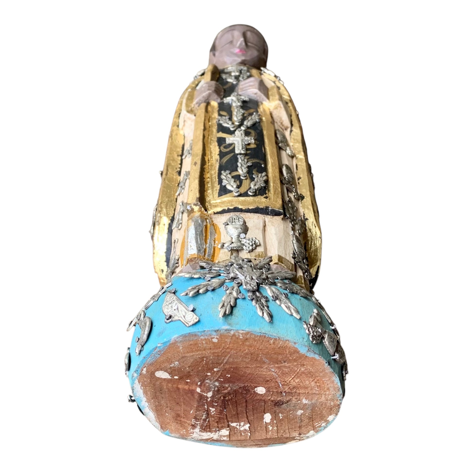 A carved milagros religious figurine of  Saint Martin in ornate black and gold vestments with silver cross details, photographed outdoors in front of a blurred stone archway. Bottom view of the wood figurine.