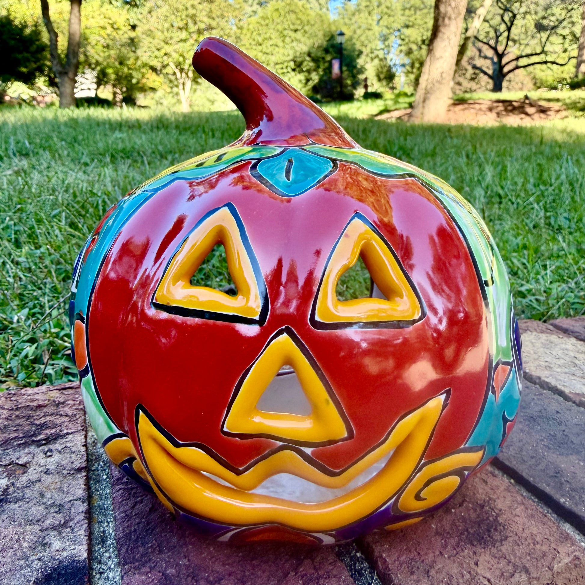 A glossy talavera jack-o'-lantern pumpkin decoration with a red face and multi-color designs sitting outdoors on a brick surface with a green grassy lawn and trees in the background.