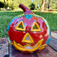 A glossy talavera jack-o'-lantern pumpkin decoration with a red face and multi-color designs sitting outdoors on a brick surface with a green grassy lawn and trees in the background.