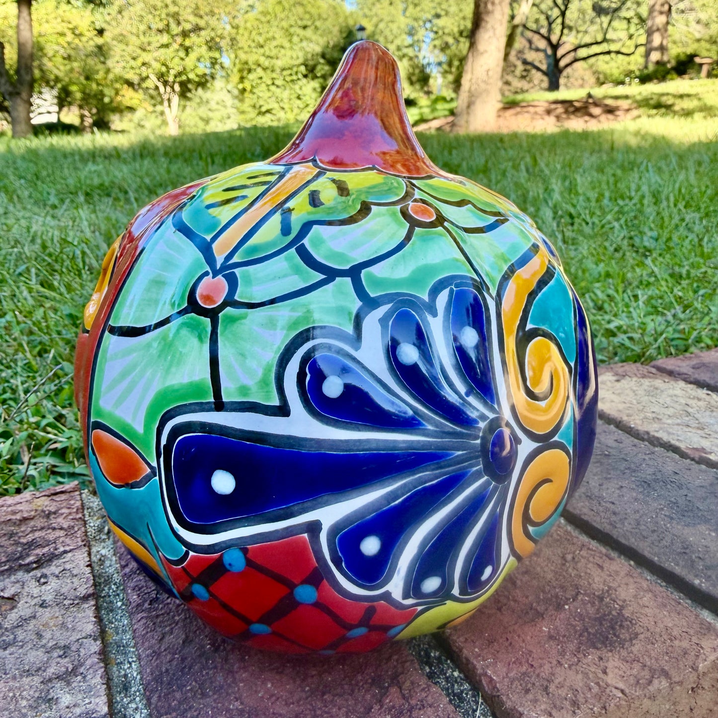 A glossy talavera jack-o'-lantern pumpkin decoration with a red face and multi-color designs sitting outdoors on a brick surface with a green grassy lawn and trees in the background. Right side view of the pumpkin.