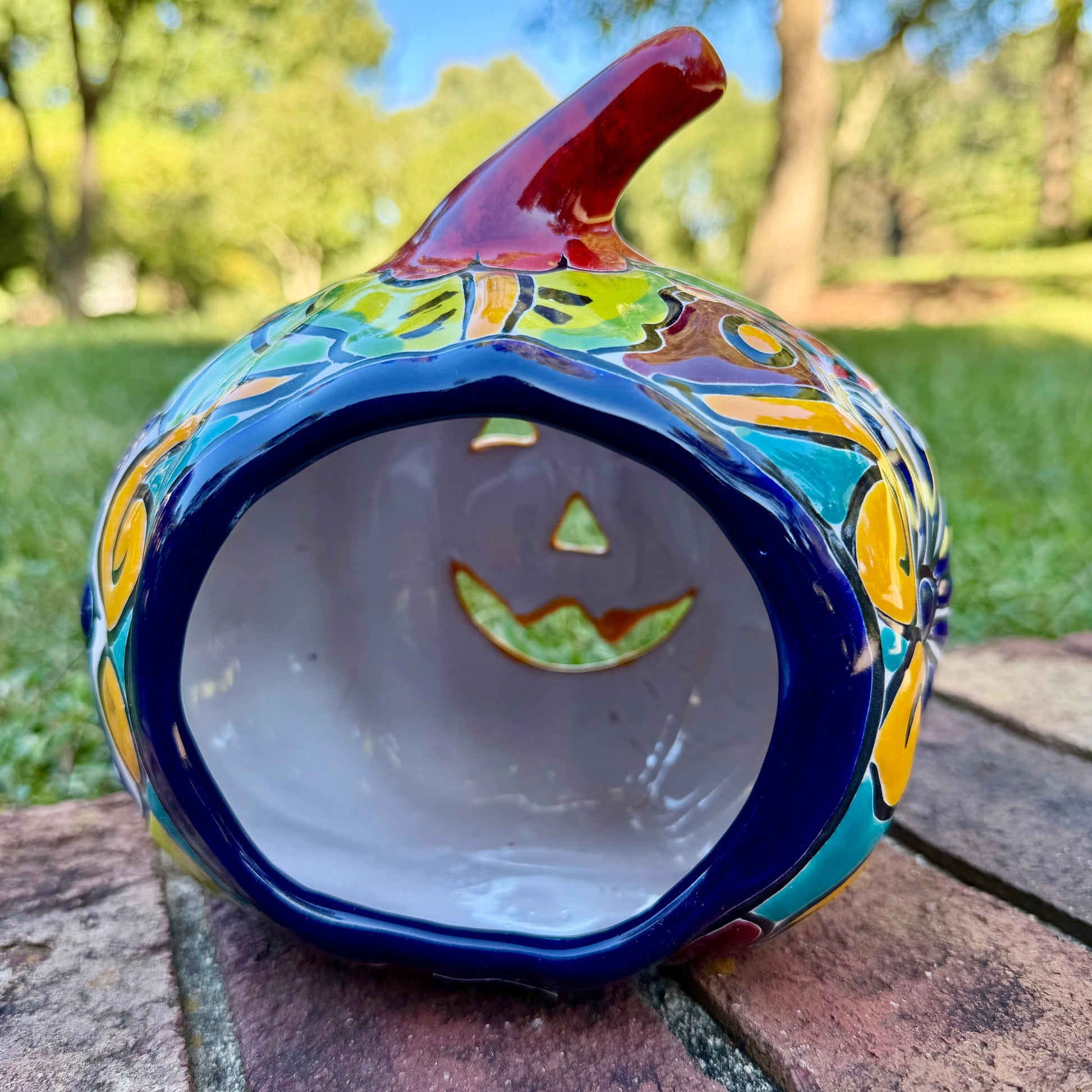 A glossy talavera jack-o'-lantern pumpkin decoration with a red face and multi-color designs sitting outdoors on a brick surface with a green grassy lawn and trees in the background. This is the back view.