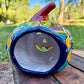 A glossy talavera jack-o'-lantern pumpkin decoration with a red face and multi-color designs sitting outdoors on a brick surface with a green grassy lawn and trees in the background. This is the back view.