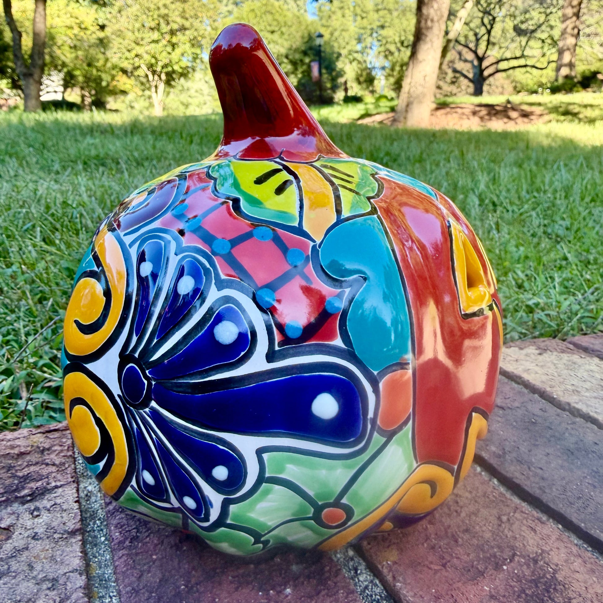 A glossy talavera jack-o'-lantern pumpkin decoration with a red face and multi-color designs sitting outdoors on a brick surface with a green grassy lawn and trees in the background. This is the left side view.
