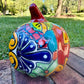 A glossy talavera jack-o'-lantern pumpkin decoration with a red face and multi-color designs sitting outdoors on a brick surface with a green grassy lawn and trees in the background. This is the left side view.