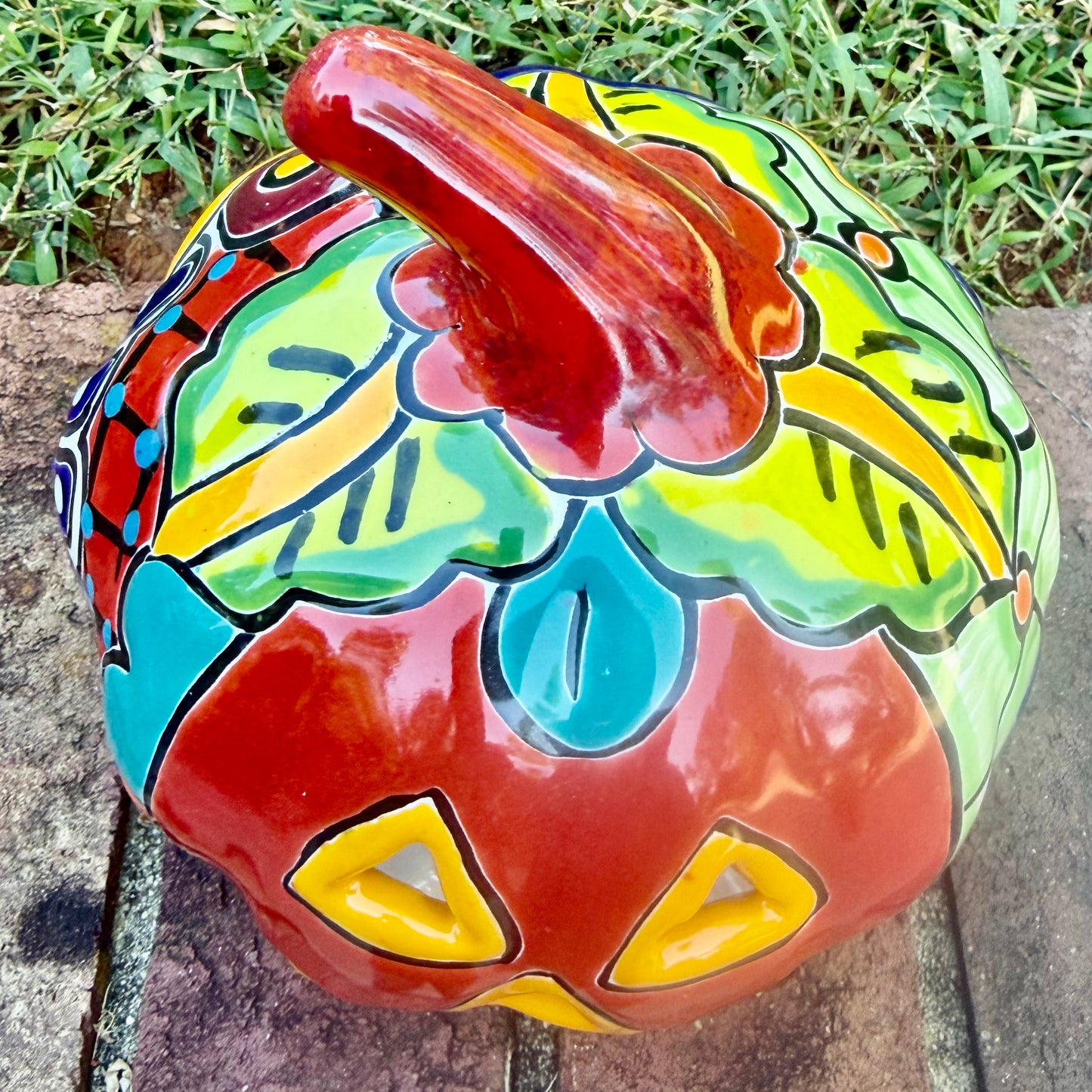 A glossy talavera jack-o'-lantern pumpkin decoration with a red face and multi-color designs sitting outdoors on a brick surface with a green grassy lawn and trees in the background. This is the top view.