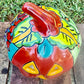A glossy talavera jack-o'-lantern pumpkin decoration with a red face and multi-color designs sitting outdoors on a brick surface with a green grassy lawn and trees in the background. This is the top view.