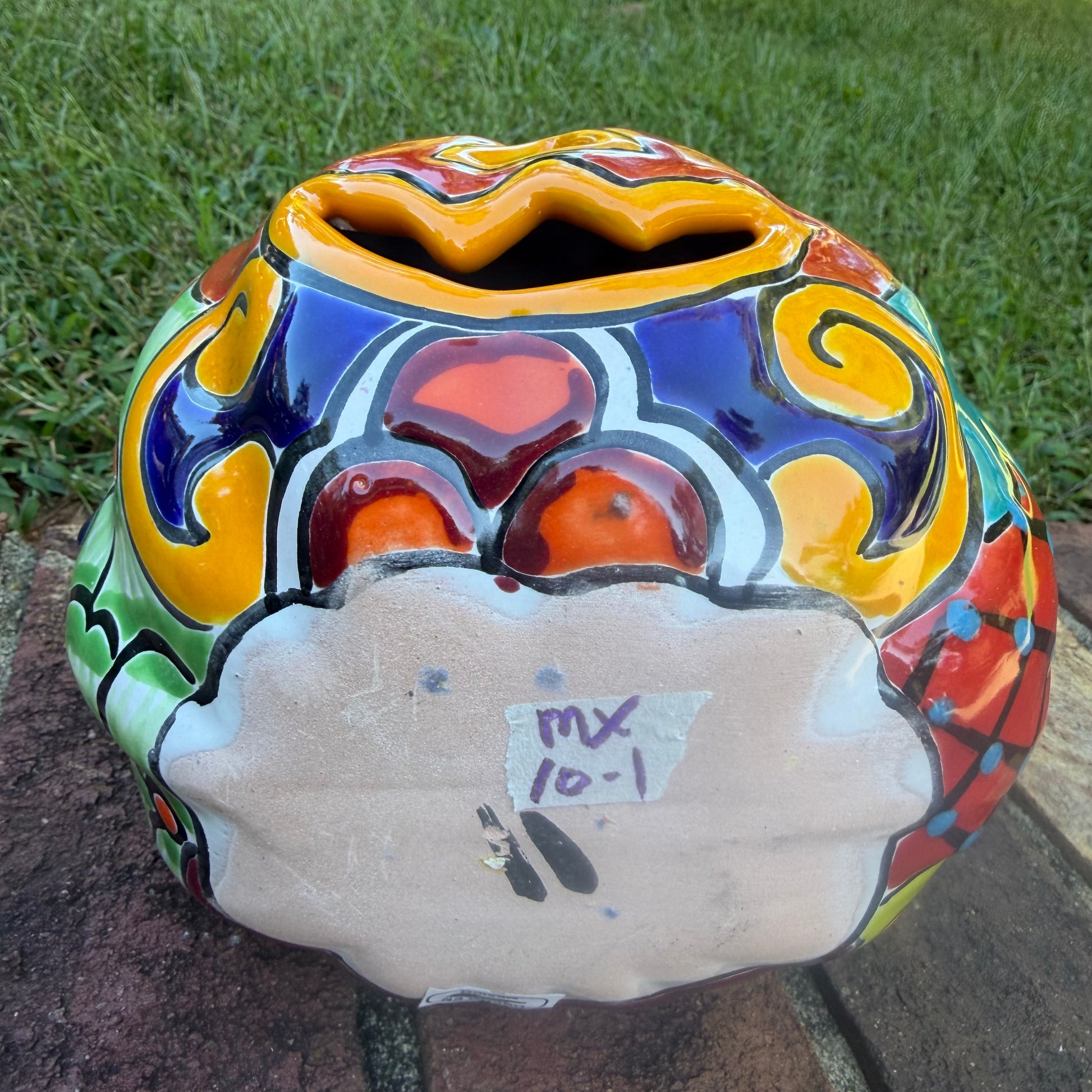 A glossy talavera jack-o'-lantern pumpkin decoration with a red face and multi-color designs sitting outdoors on a brick surface with a green grassy lawn and trees in the background. This is the bottom view.