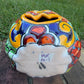 A glossy talavera jack-o'-lantern pumpkin decoration with a red face and multi-color designs sitting outdoors on a brick surface with a green grassy lawn and trees in the background. This is the bottom view.