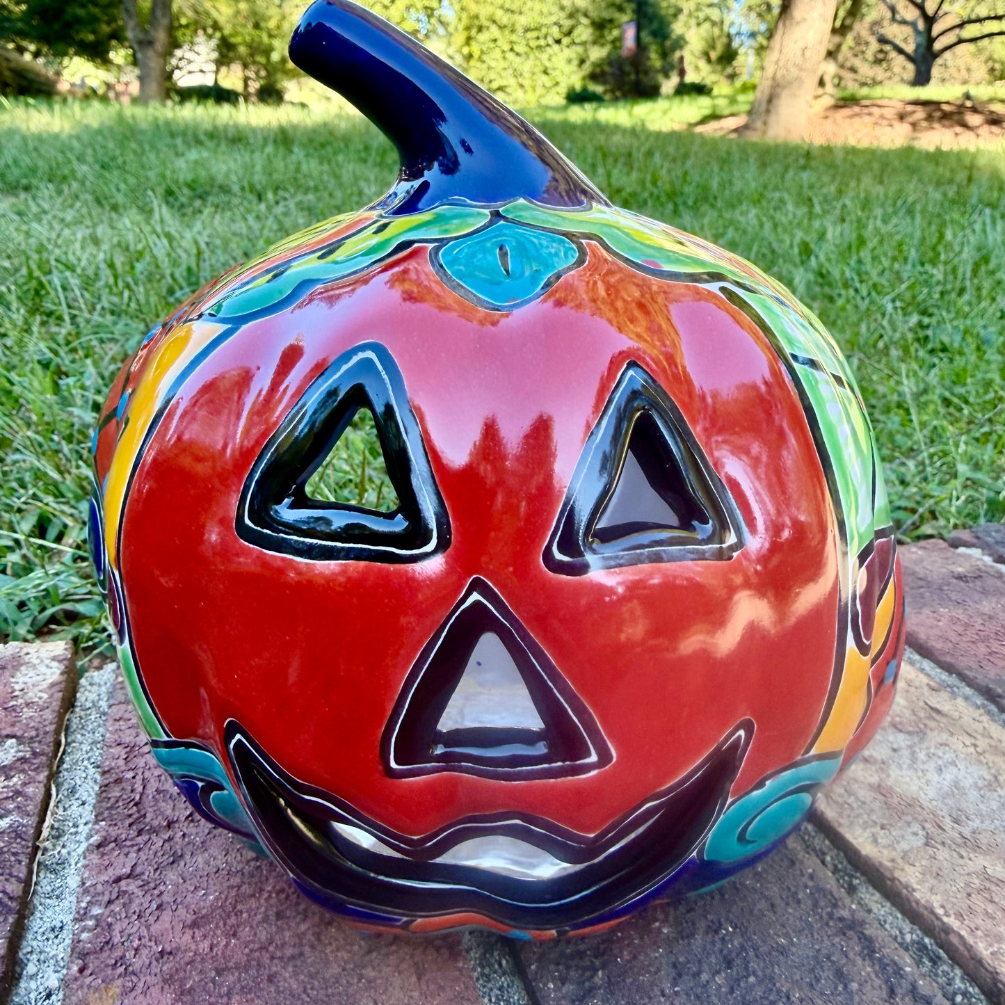 A glossy talavera jack-o'-lantern pumpkin with a red face and hand-painted multi-color designs, sitting on a brick path in a grassy outdoor yard.