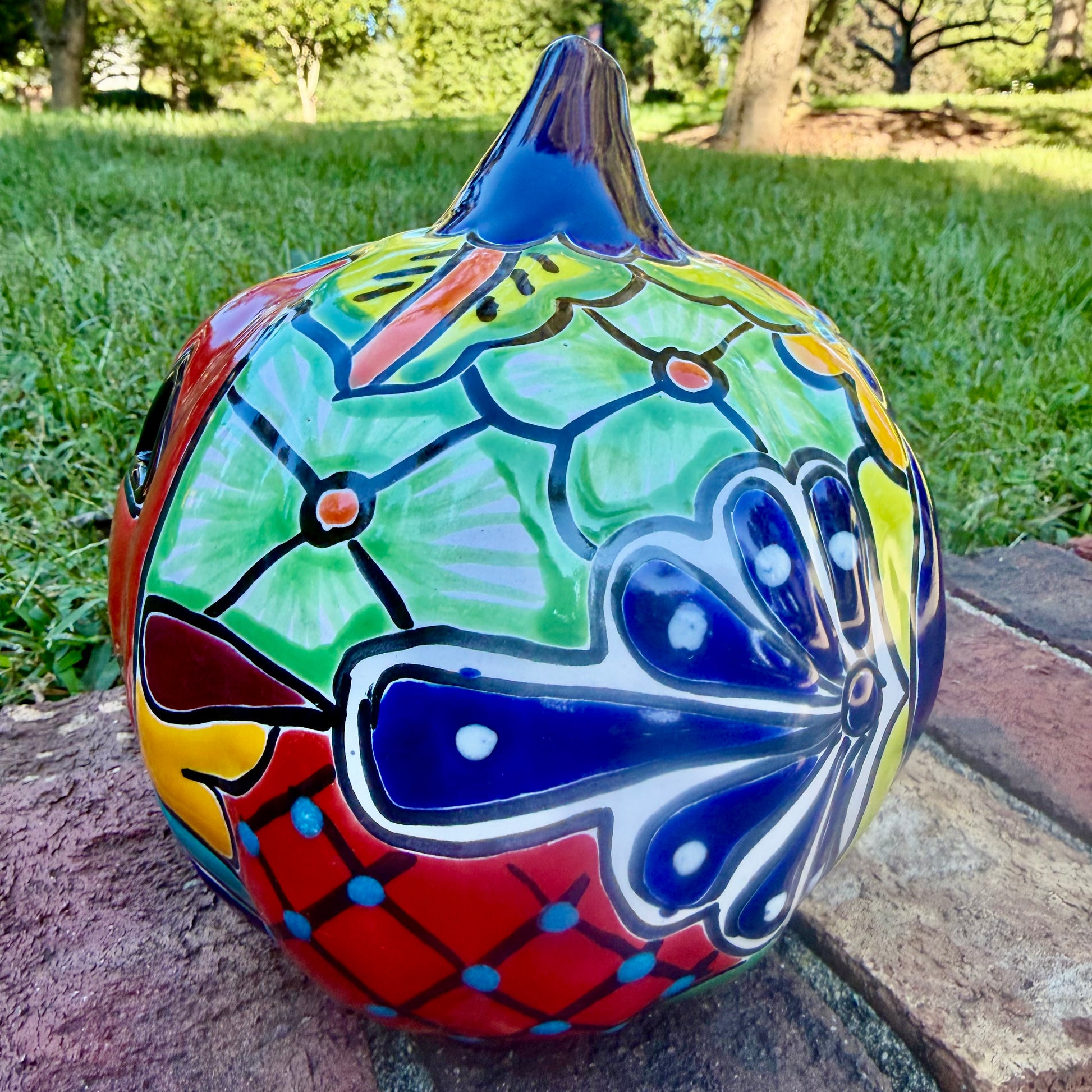 A glossy talavera jack-o'-lantern pumpkin with a red face and hand-painted multi-color designs, sitting on a brick path in a grassy outdoor yard. this is the right side view.