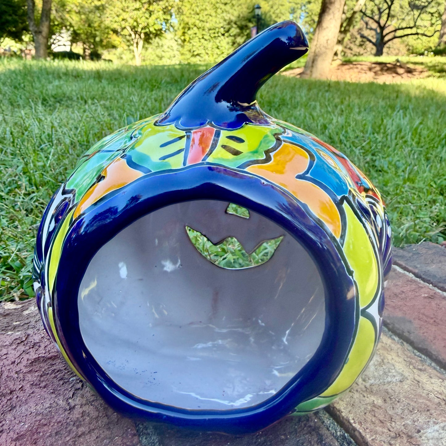 A glossy talavera jack-o'-lantern pumpkin with a red face and hand-painted multi-color designs, sitting on a brick path in a grassy outdoor yard. This is the back view showing candle opening.
