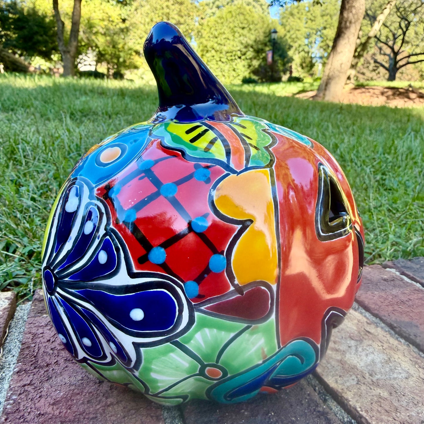 A glossy talavera jack-o'-lantern pumpkin with a red face and hand-painted multi-color designs, sitting on a brick path in a grassy outdoor yard. This is the left side view.