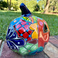 A glossy talavera jack-o'-lantern pumpkin with a red face and hand-painted multi-color designs, sitting on a brick path in a grassy outdoor yard. This is the left side view.