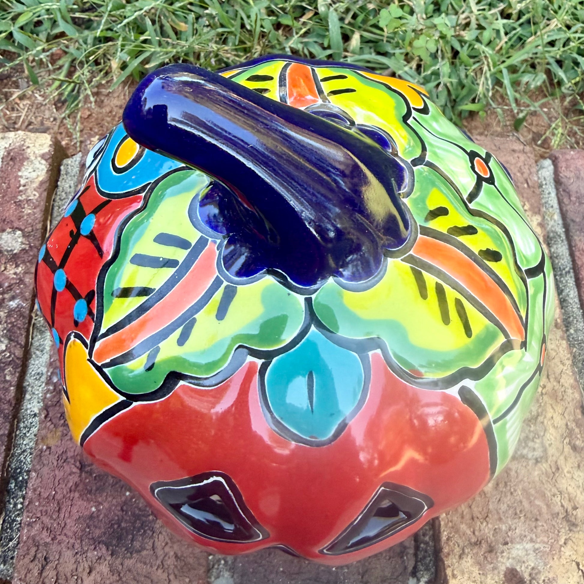 A glossy talavera jack-o'-lantern pumpkin with a red face and hand-painted multi-color designs, sitting on a brick path in a grassy outdoor yard. This is the top view of the stem.