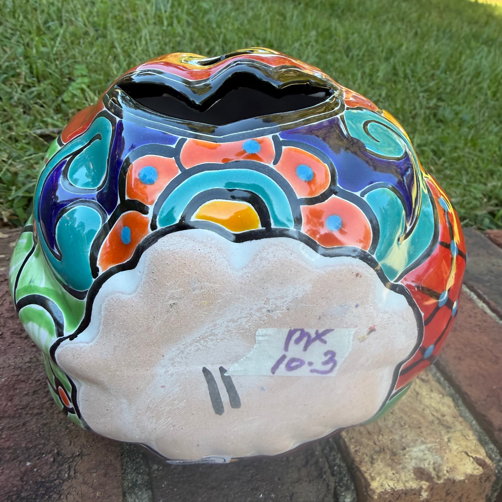 A glossy talavera jack-o'-lantern pumpkin with a red face and hand-painted multi-color designs, sitting on a brick path in a grassy outdoor yard. This is the bottom view.