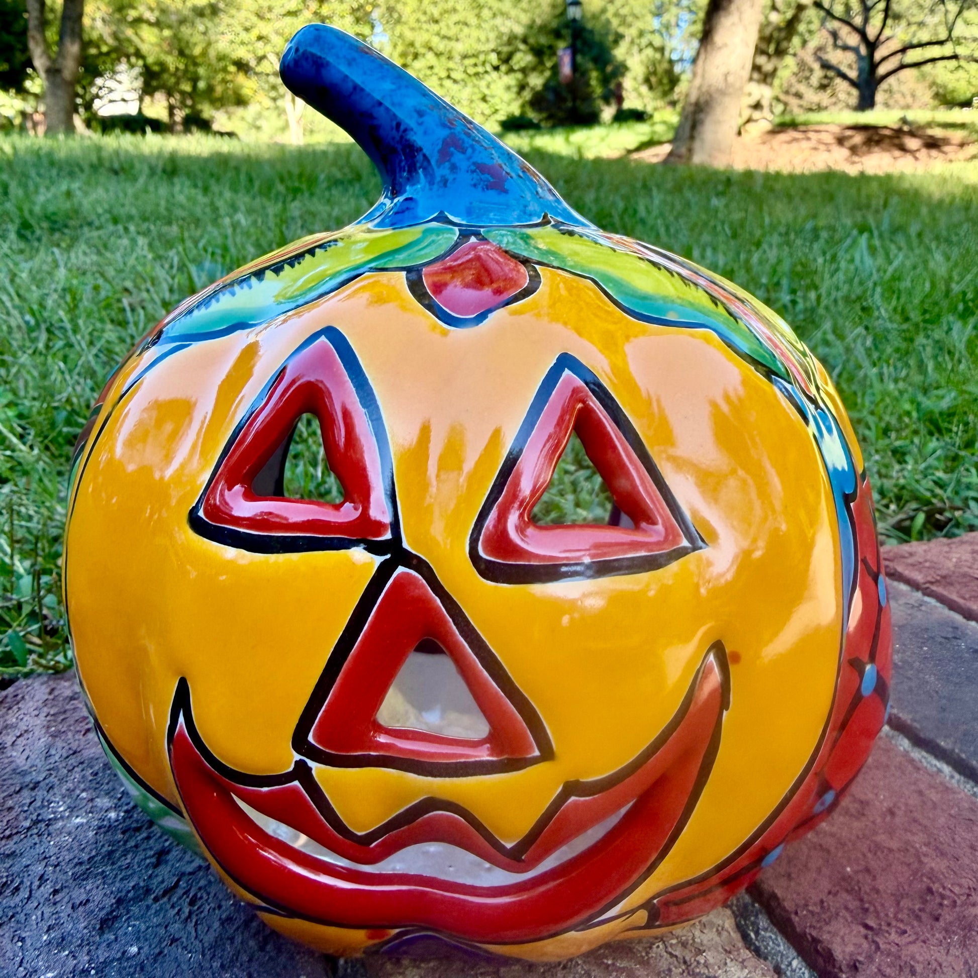 A hand-painted talavera jack-o'-lantern pumpkin garden ornament sitting outdoors on a brick path with green grass and trees in the background. The pumpkin features a bright yellow face with red and black painted facial features, a blue stem, and multicolored painted accents on top.