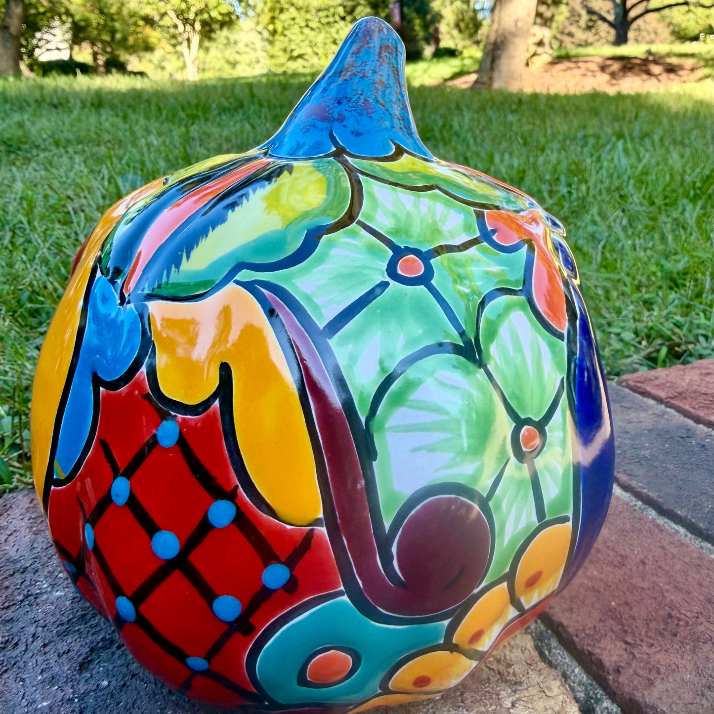 A hand-painted talavera jack-o'-lantern pumpkin garden ornament sitting outdoors on a brick path with green grass and trees in the background. The pumpkin features a bright yellow face with red and black painted facial features, a blue stem, and multicolored painted accents on top.  This is the right side view.