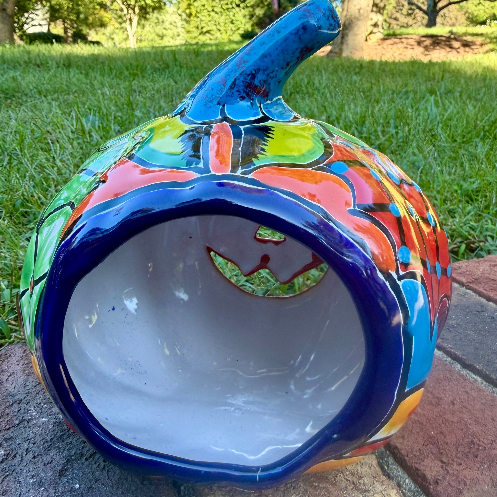 A hand-painted talavera jack-o'-lantern pumpkin garden ornament sitting outdoors on a brick path with green grass and trees in the background. The pumpkin features a bright yellow face with red and black painted facial features, a blue stem, and multicolored painted accents on top. This is the back view showing the candle opening.