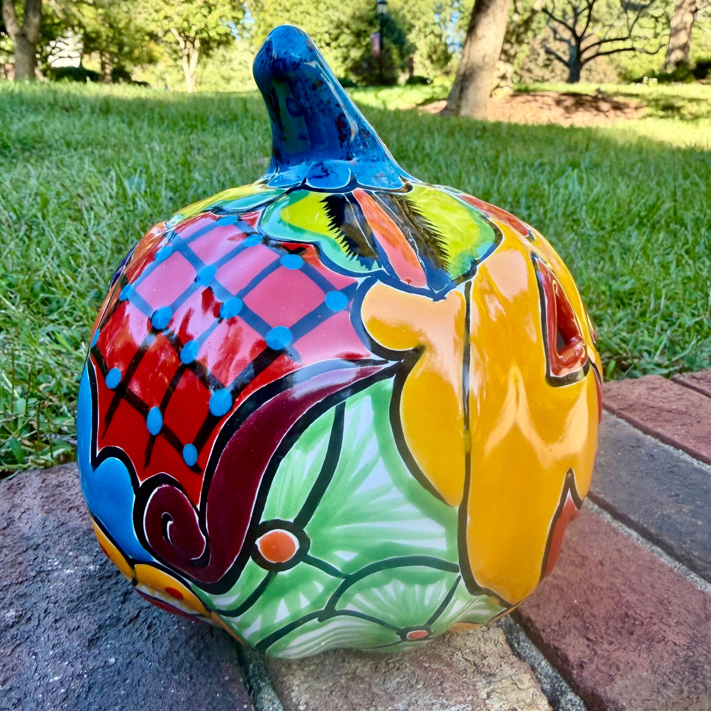 A hand-painted talavera jack-o'-lantern pumpkin garden ornament sitting outdoors on a brick path with green grass and trees in the background. The pumpkin features a bright yellow face with red and black painted facial features, a blue stem, and multicolored painted accents on top. This is the left side view.