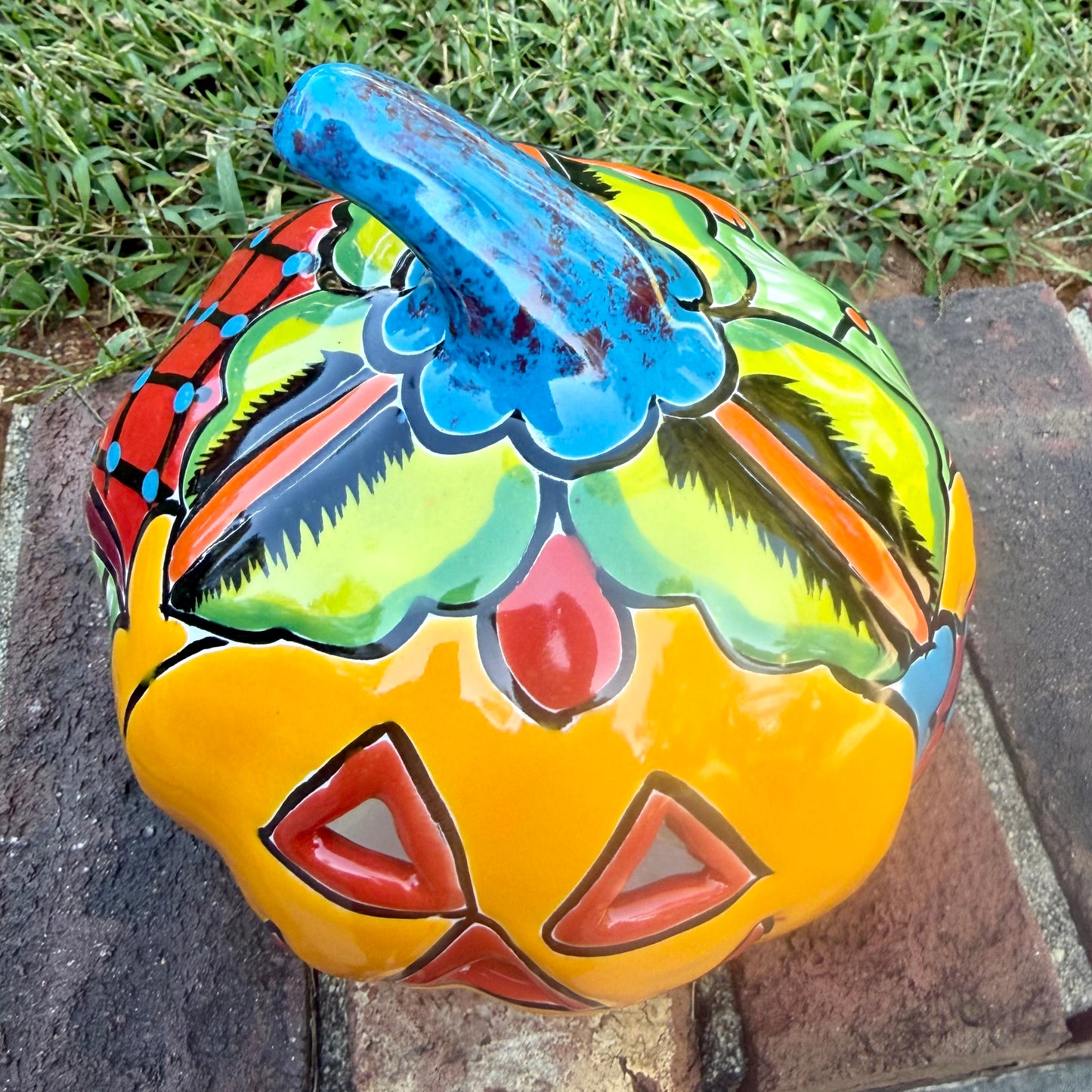 A hand-painted talavera jack-o'-lantern pumpkin garden ornament sitting outdoors on a brick path with green grass and trees in the background. The pumpkin features a bright yellow face with red and black painted facial features, a blue stem, and multicolored painted accents on top. This is the top view.