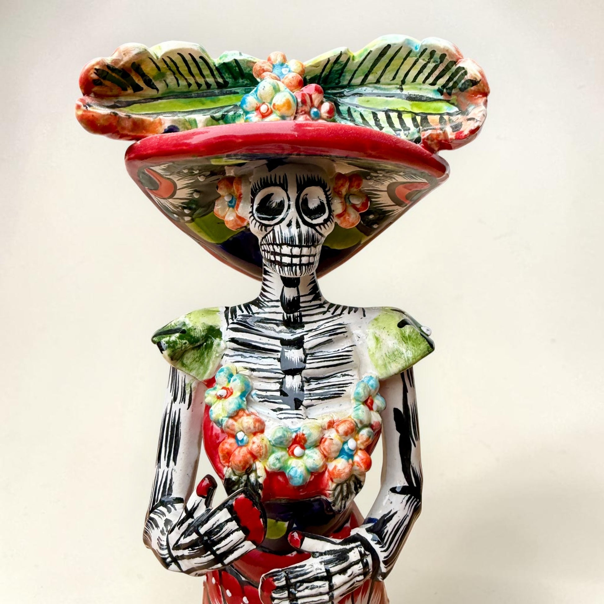 A colorful hand-painted talavera Day of the Dead (Día de los Muertos) skeleton figurine wearing a wide-brimmed sombrero and a floral dress, set against a plain off-white background. This is a photo of the face close up.