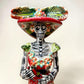 A colorful hand-painted talavera Day of the Dead (Día de los Muertos) skeleton figurine wearing a wide-brimmed sombrero and a floral dress, set against a plain off-white background. This is a photo of the face close up.