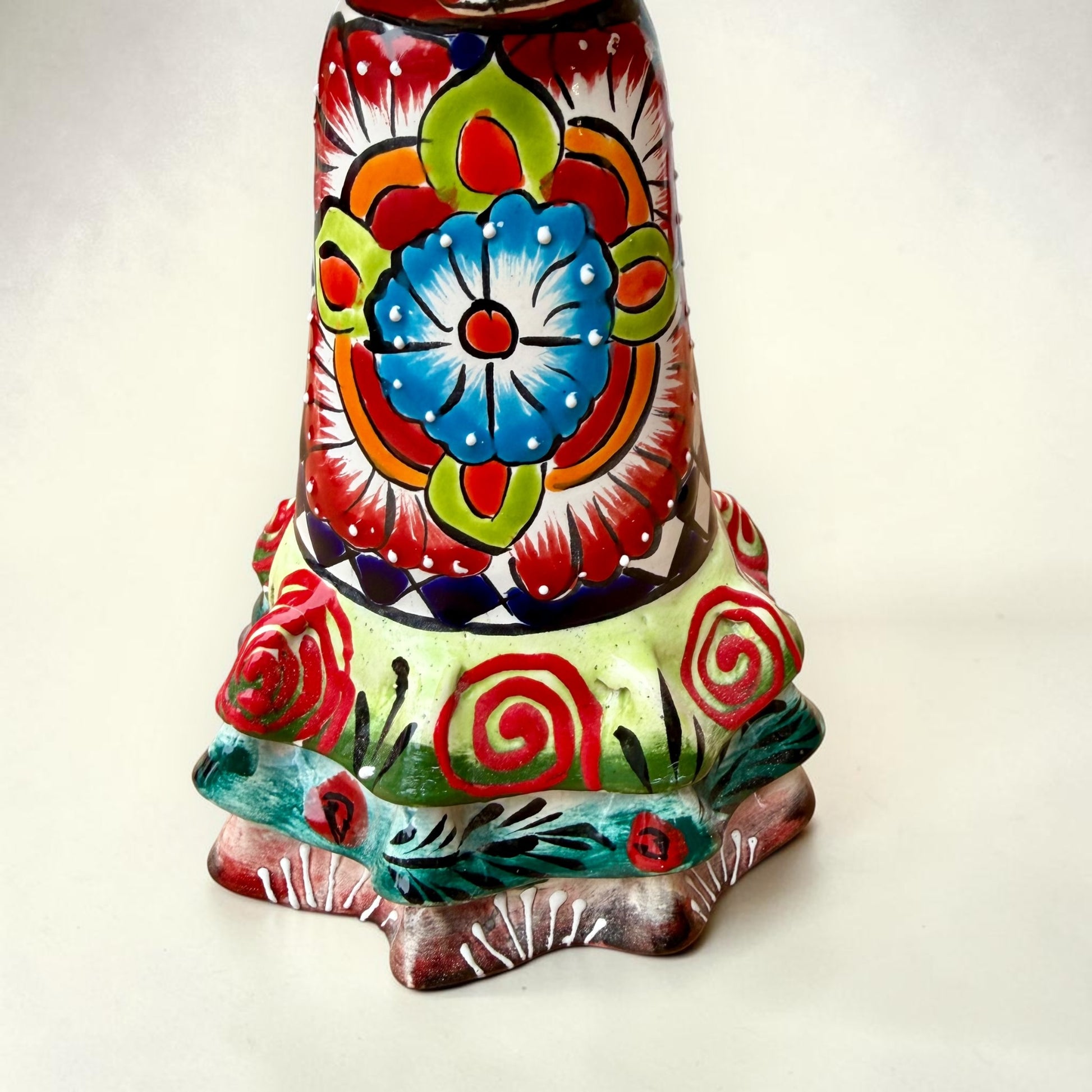 A colorful hand-painted talavera Day of the Dead (Día de los Muertos) skeleton figurine wearing a wide-brimmed sombrero and a floral dress, set against a plain off-white background. This is a photo of the ruffle at the bottom of her dress.