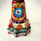 A colorful hand-painted talavera Day of the Dead (Día de los Muertos) skeleton figurine wearing a wide-brimmed sombrero and a floral dress, set against a plain off-white background. This is a photo of the ruffle at the bottom of her dress.