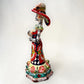 A colorful hand-painted talavera Day of the Dead (Día de los Muertos) skeleton figurine wearing a wide-brimmed sombrero and a floral dress, set against a plain off-white background. right side view of the figurine.