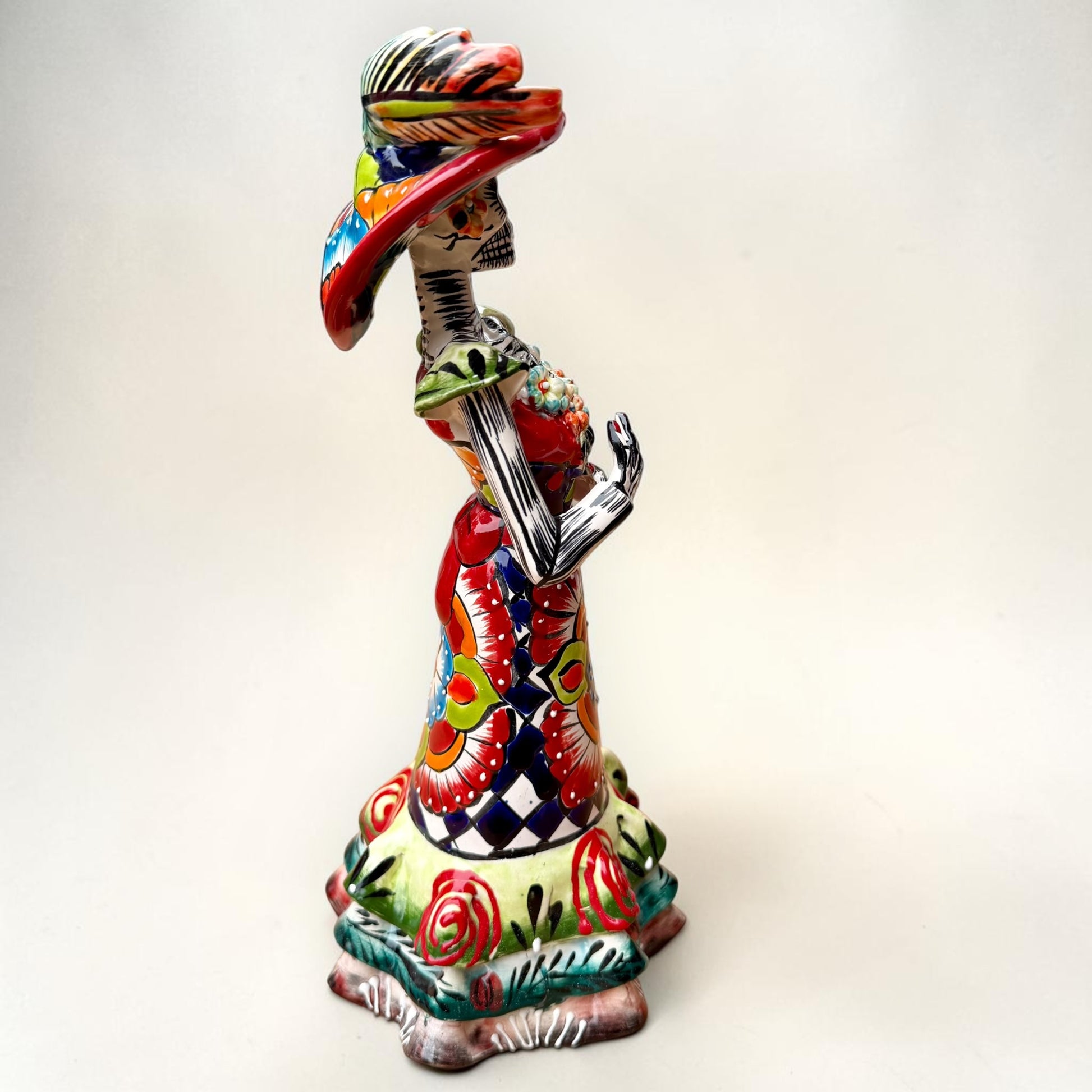 A colorful hand-painted talavera Day of the Dead (Día de los Muertos) skeleton figurine wearing a wide-brimmed sombrero and a floral dress, set against a plain off-white background. left side view of the catrina doll.