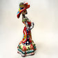 A colorful hand-painted talavera Day of the Dead (Día de los Muertos) skeleton figurine wearing a wide-brimmed sombrero and a floral dress, set against a plain off-white background. left side view of the catrina doll.