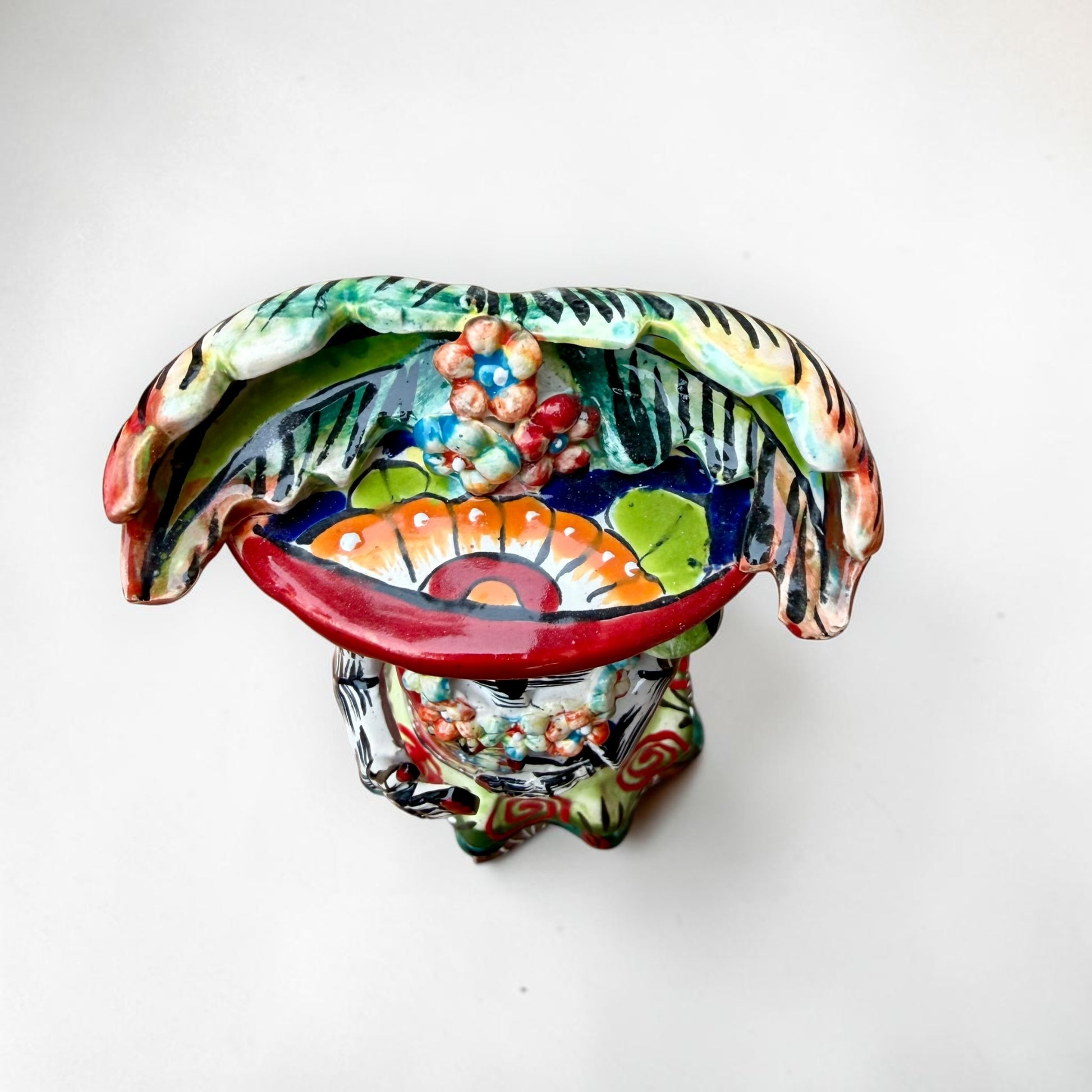 A colorful hand-painted talavera Day of the Dead (Día de los Muertos) skeleton figurine wearing a wide-brimmed sombrero and a floral dress, set against a plain off-white background. This is a view from the top of the doll.