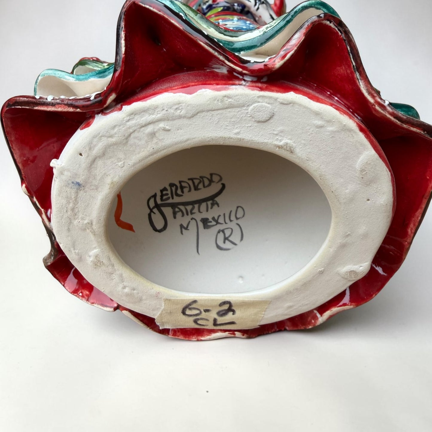 A colorful hand-painted talavera Day of the Dead (Día de los Muertos) skeleton figurine wearing a wide-brimmed sombrero and a floral dress, set against a plain off-white background.  Bottom of the doll showing artist signature.