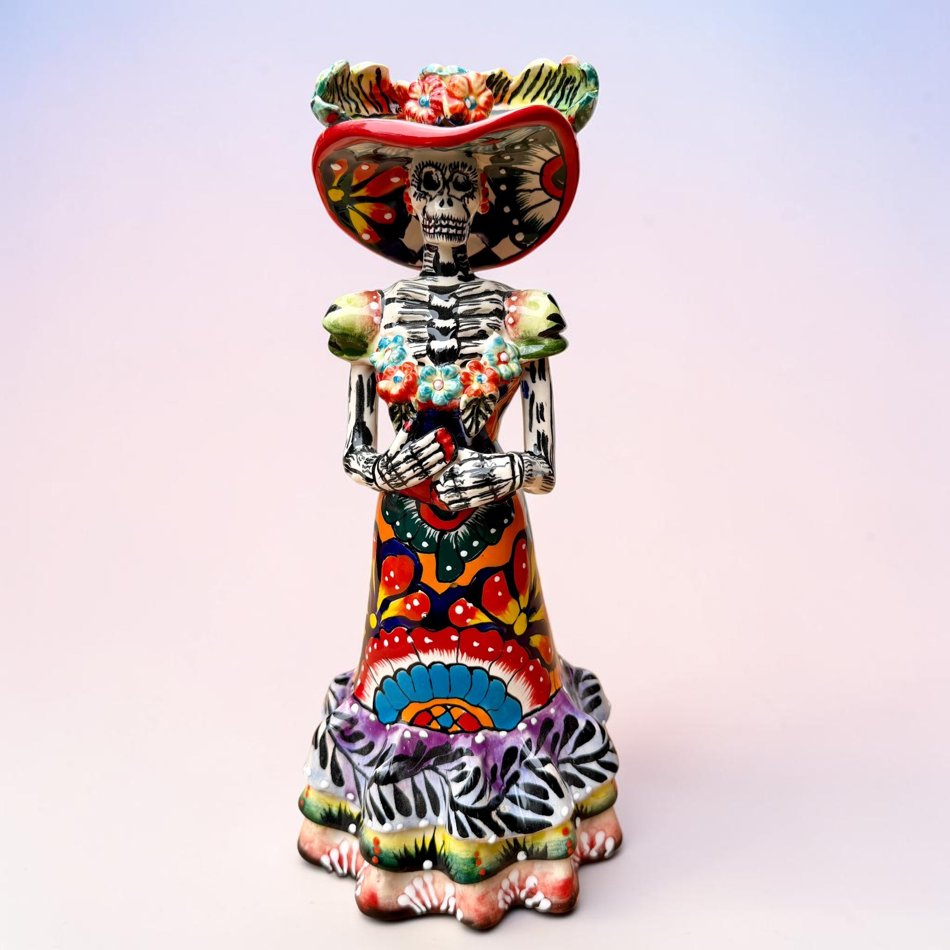 A colorful Day of the Dead talavera skeleton figurine wearing a wide-brimmed red hat and a vibrant floral dress, set against a soft light pink gradient background.