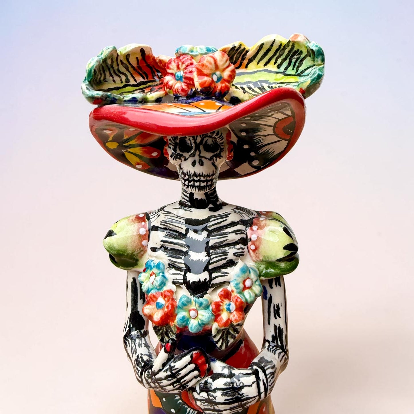 A colorful Day of the Dead talavera skeleton figurine wearing a wide-brimmed red hat and a vibrant floral dress, set against a soft light pink gradient background. Close up of face.
