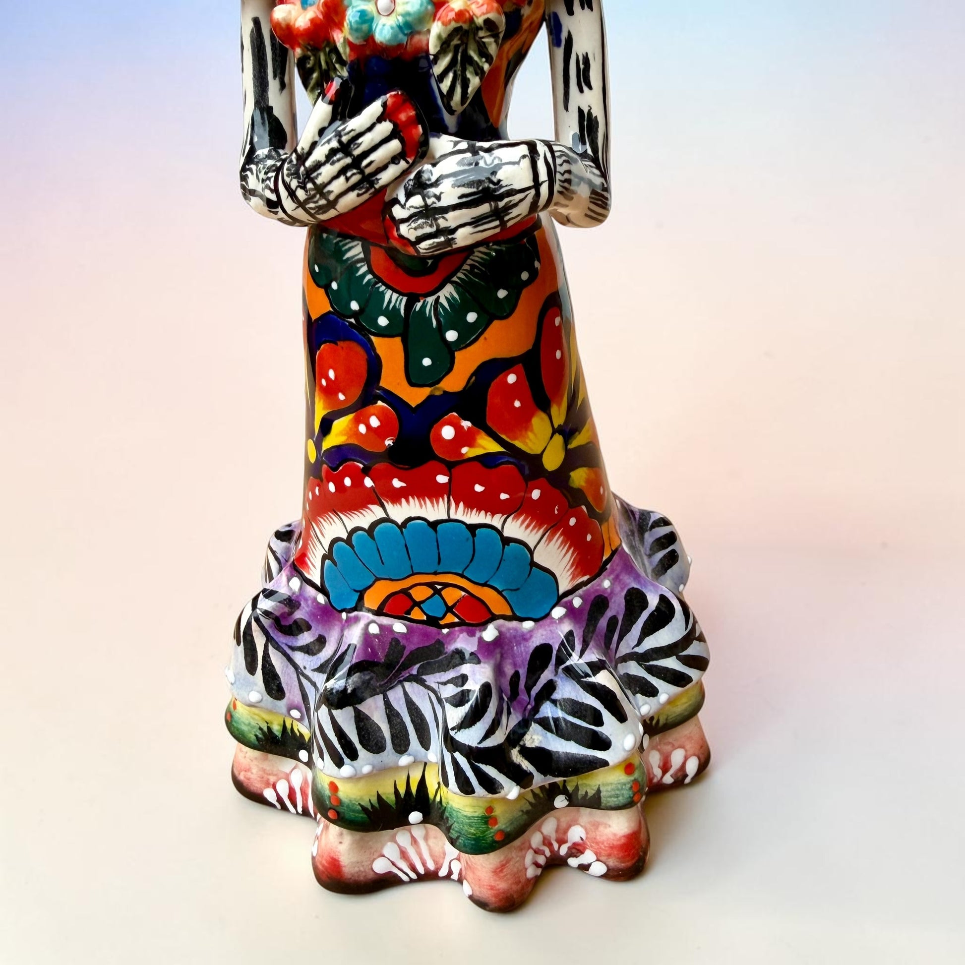 A colorful Day of the Dead talavera skeleton figurine wearing a wide-brimmed red hat and a vibrant floral dress, set against a soft light pink gradient background.  Photo of ruffle on the bottom of her dress.