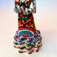 A colorful Day of the Dead talavera skeleton figurine wearing a wide-brimmed red hat and a vibrant floral dress, set against a soft light pink gradient background.  Photo of ruffle on the bottom of her dress.