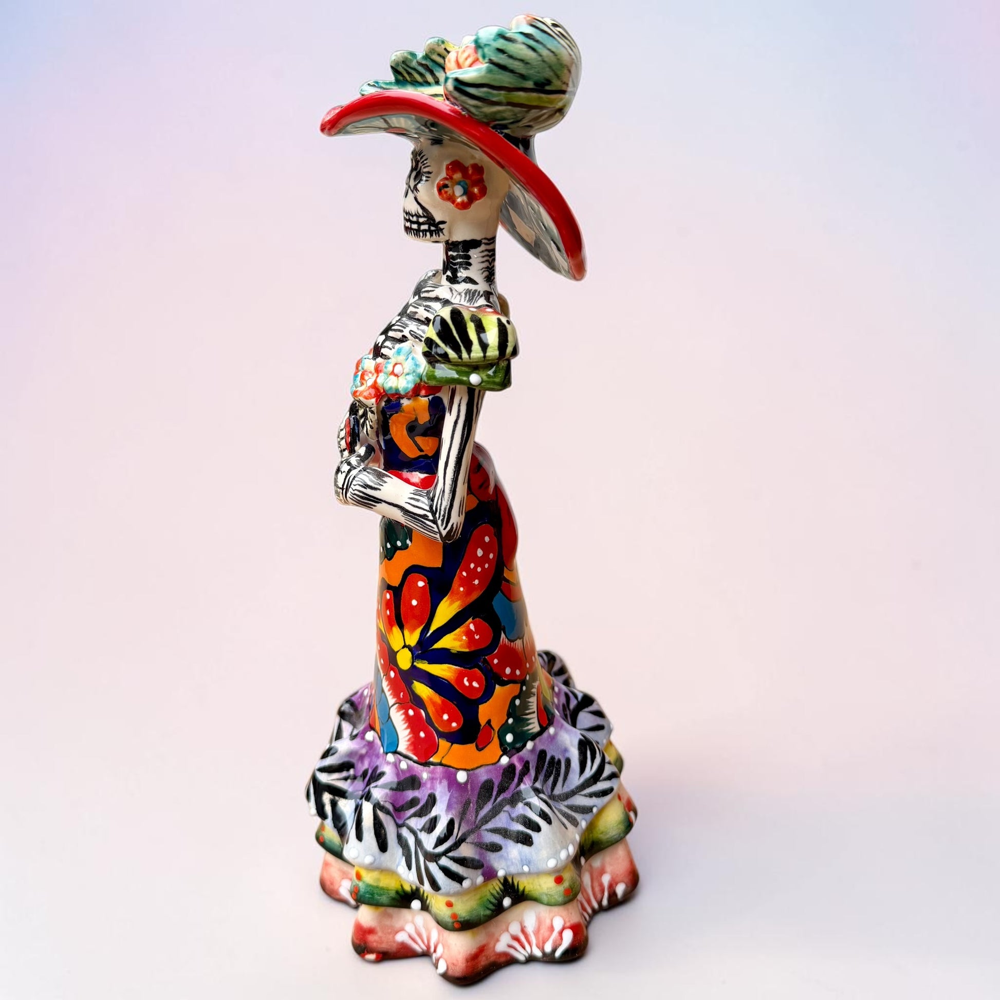 A colorful Day of the Dead talavera skeleton figurine wearing a wide-brimmed red hat and a vibrant floral dress, set against a soft light pink gradient background. Left side view of the doll.
