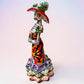 A colorful Day of the Dead talavera skeleton figurine wearing a wide-brimmed red hat and a vibrant floral dress, set against a soft light pink gradient background. Left side view of the doll.