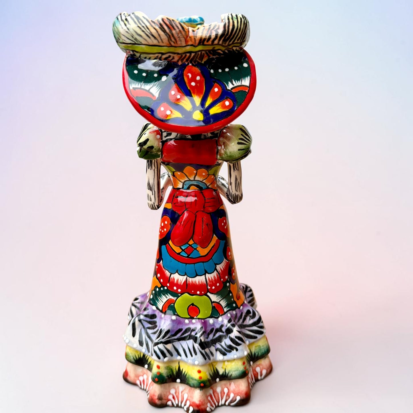 A colorful Day of the Dead talavera skeleton figurine wearing a wide-brimmed red hat and a vibrant floral dress, set against a soft light pink gradient background. Back view of doll showing the bow around her waist.