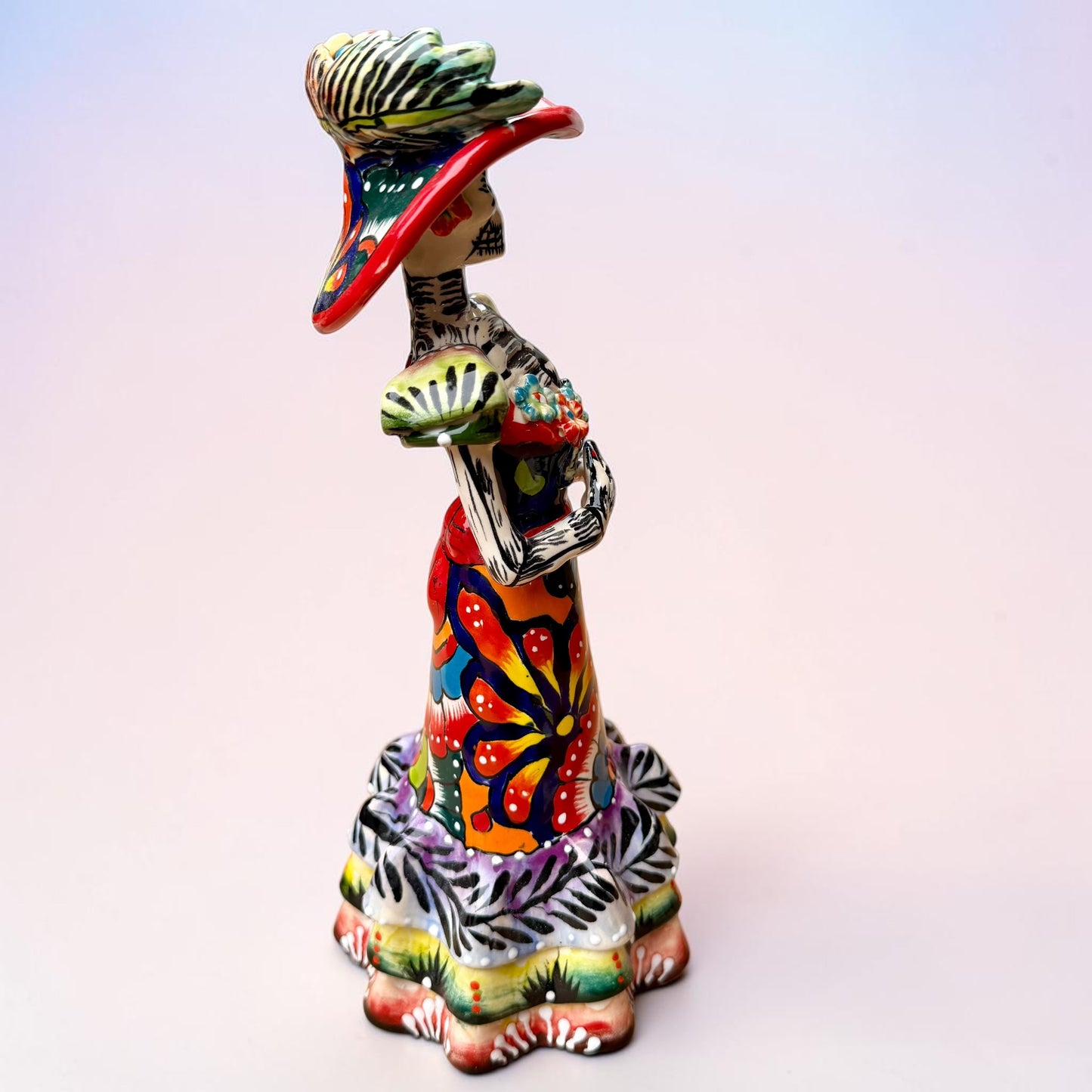 A colorful Day of the Dead talavera skeleton figurine wearing a wide-brimmed red hat and a vibrant floral dress, set against a soft light pink gradient background. Left side view of the catrina foll.