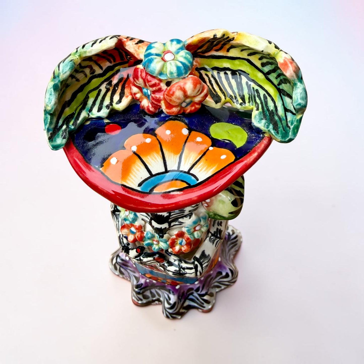 A colorful Day of the Dead talavera skeleton figurine wearing a wide-brimmed red hat and a vibrant floral dress, set against a soft light pink gradient background. Top view of the doll.